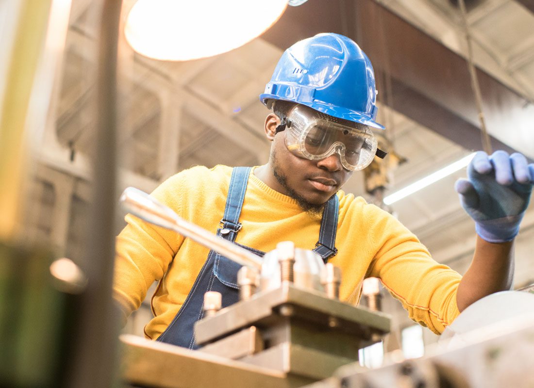 Manufacturer Insurance - Manufacturing Engineer Working in a Warehouse in a Blue Jumpsuit with a White Hard Hat and Operating a Machine